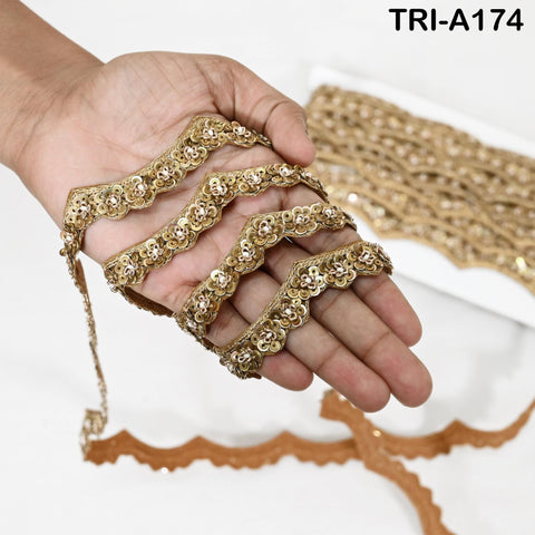 May include: Gold sequin trim with a scalloped edge and a floral design. The trim is held in a hand, highlighting its intricate details and texture. The text "TRI-A174" is visible. Suitable for craft and sewing projects. The trim is measured in centimetres.