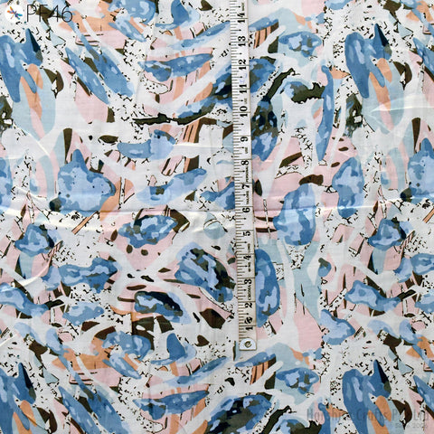 The image is a close-up of a fabric with a colorful abstract pattern, featuring various shades of blue, pink, and orange. A ruler is placed vertically in the center of the fabric, providing a reference for the size of the pattern.