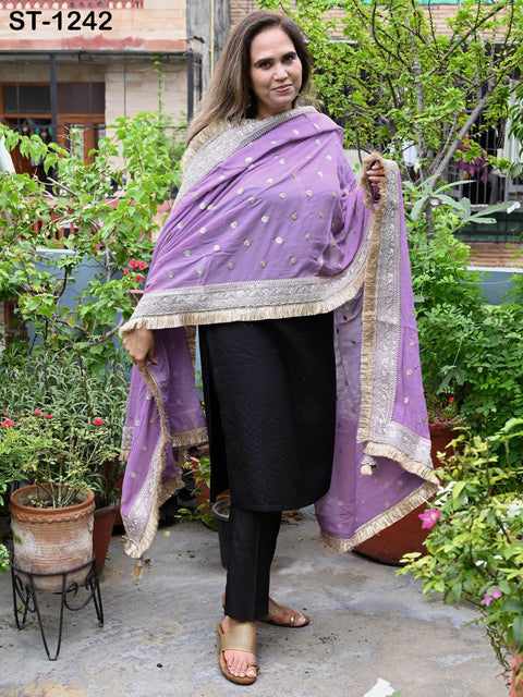 A woman wearing a purple and gold shawl stands in a garden surrounded by potted plants.