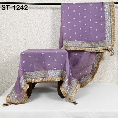 a purple and gold decorative textile, possibly a sari or a shawl, with a floral pattern and fringed edges. The textile is hanging on a rack, and there is a small table or stool in front of it. The background is a plain white wall with a floral pattern.