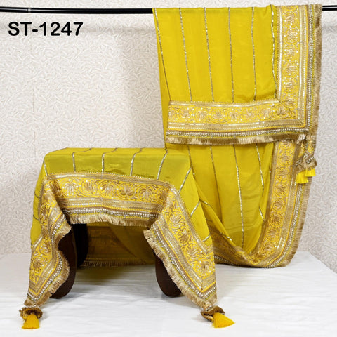a yellow and gold decorative textile, possibly a tablecloth or a garment, with intricate gold embroidery and tassels. The textile is displayed on a white background, and there is a small table or stool in front of it.