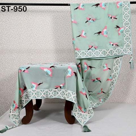 a tablecloth with a repeating pattern of pink and green birds on a light green background. The tablecloth is hanging on a wooden stand, and there is a small tablecloth placed on the floor next to it.