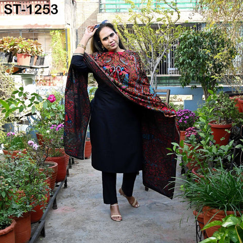 A woman in a black dress with a colorful pattern stands in a garden surrounded by potted plants.