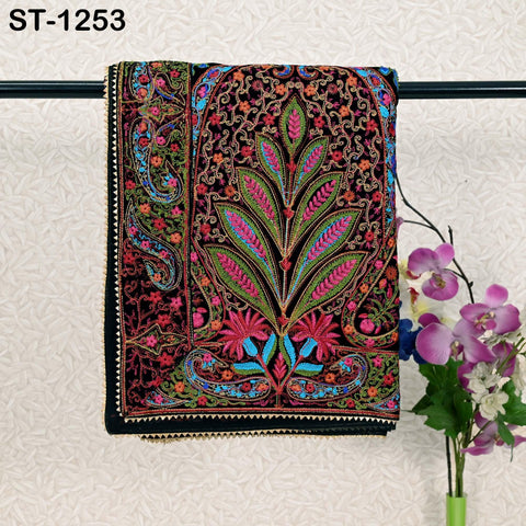 a colorful, intricately embroidered textile hanging on a black rod, with a vase of purple flowers placed next to it.