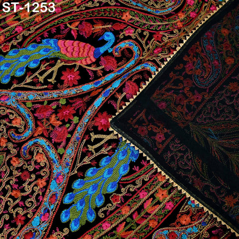 The image is a close-up of a vibrant, intricately embroidered fabric with a peacock design. The peacock is depicted in shades of blue, green, and pink, and the fabric features a variety of floral and paisley patterns in red, black, and gold. The background is a dark, textured fabric with a subtle pattern.