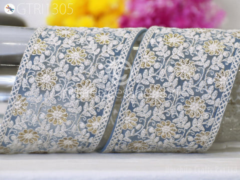 a close-up view of a decorative fabric with a floral pattern, including white and gold flowers. The fabric appears to be a decorative ribbon or lace, with a repeating floral design. The background is blurred, but it appears to be a colorful floral arrangement.