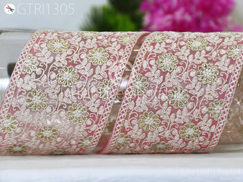 a close-up view of a decorative fabric with a floral pattern in shades of pink and white. The fabric appears to be a ribbon or lace, with intricate embroidered designs. The background is blurred, but it appears to be a floral arrangement, adding to the overall aesthetic of the image.