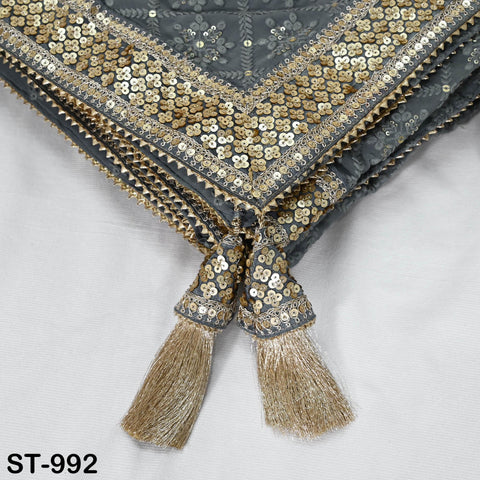 a close-up view of a decorative fabric with a gold and black sequined trim. The fabric has a tassel attached to it, which is made of gold and has a tassel fringe. The background is a plain white fabric.