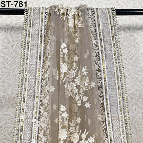 a close-up view of a decorative fabric with intricate floral patterns and a sheer, lace-like texture. The fabric appears to be a sheer lace or chiffon material, with a floral design and beaded or sequined trim along the edges. The fabric is hanging on a black rod or stand, which is placed against a textured background.