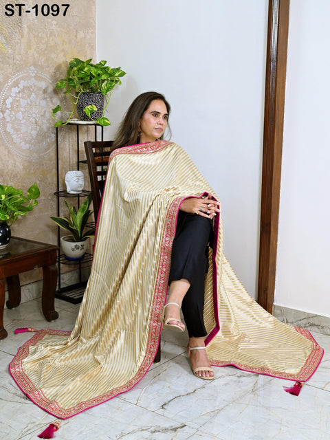 A woman wearing a long, beige and pink striped shawl is sitting on a chair in a room with a potted plant and a wooden shelf.
