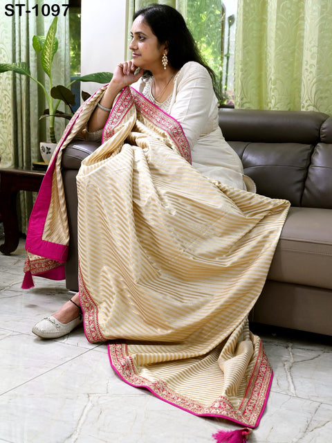 A woman in a white dress sits on a couch, wearing a pink and gold sari with a matching pink and gold shawl draped over her shoulders.