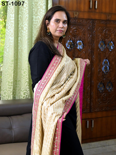 A woman wearing a black dress and a gold and white patterned shawl stands in front of a wooden cabinet.