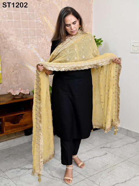 A woman wearing a black dress and a yellow and gold shawl stands in a room with a pink wall and a wooden cabinet.
