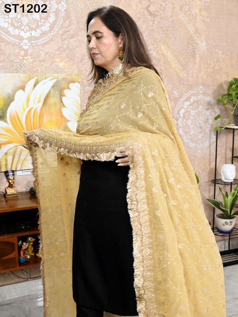 A woman wearing a yellow and gold shawl stands in a room with a floral patterned wall behind her.