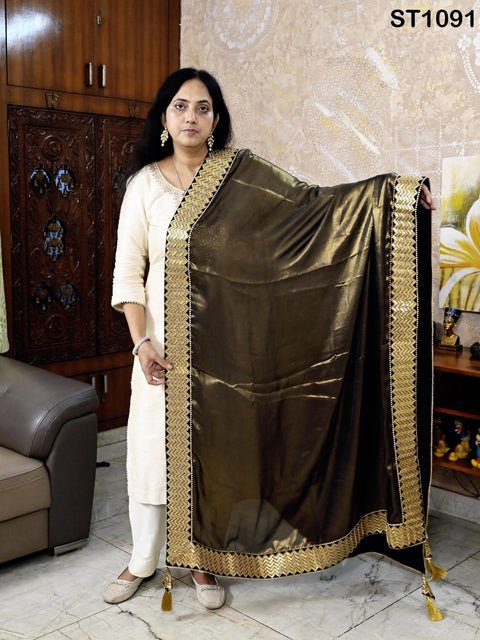 A woman in a white dress stands in a room, holding a black and gold shawl with a gold trim.