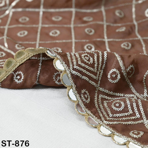 a close-up view of a brown fabric with intricate white and gold embroidery patterns, including a diamond-shaped design and circular motifs. The fabric appears to be a piece of clothing or textile with a decorative, ornamental design.