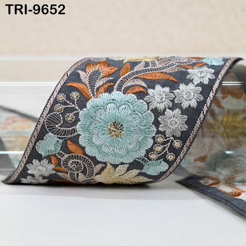 a decorative fabric with a floral pattern, including a large blue flower and smaller white flowers. The fabric appears to be a decorative trim or border, with intricate embroidered designs.