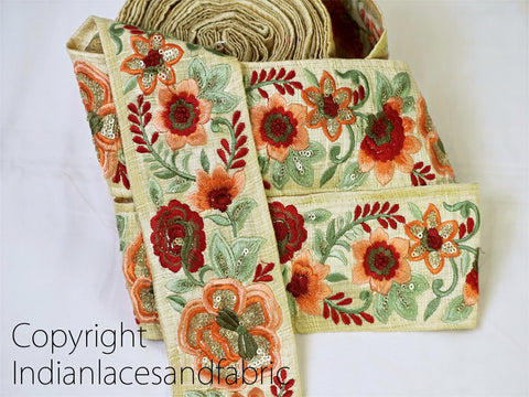 a close-up view of a decorative fabric with a floral pattern, including red, orange, and green flowers. The fabric appears to be a beige or off-white color, and the intricate embroidery details suggest a high level of craftsmanship.