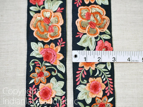 two pieces of fabric with intricate floral embroidery, including red, orange, and green flowers, as well as leaves and other decorative elements. A ruler is placed between the two pieces of fabric to measure their length.