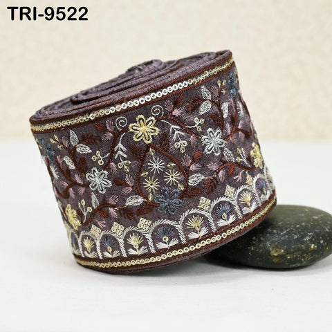 a decorative fabric with intricate floral patterns and designs, including a variety of colors such as purple, gold, and white. The fabric is rolled up and placed on a surface, with a rock visible underneath it.