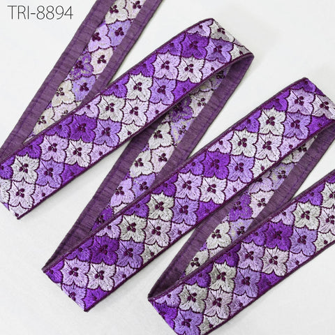 a close-up view of a decorative fabric with a repeating pattern of purple and white flowers. The fabric appears to be a decorative trim or ribbon, with a purple border and a repeating pattern of purple and white flowers. The fabric is neatly arranged, and the image does not contain any text or other objects.
