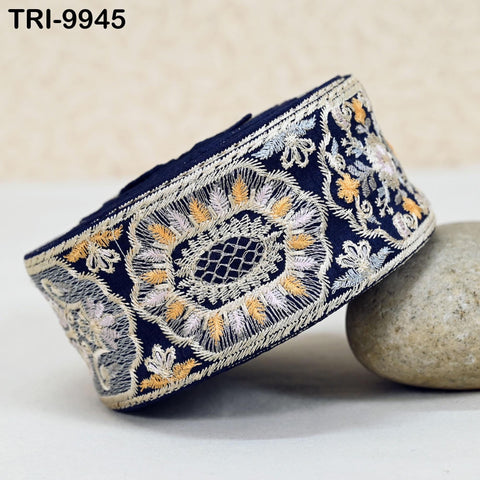 a decorative blue and white fabric with intricate embroidered patterns, including a large floral design and smaller floral and leaf motifs. The fabric is displayed on a white surface, with a small rock placed next to it.