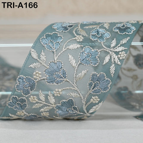a close-up view of a decorative fabric with a floral pattern. The fabric appears to be a light blue color, and the pattern consists of blue flowers and leaves with white accents. The fabric is folded and displayed on a surface, possibly a table or a shelf.