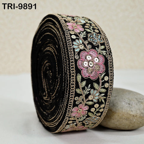 A close-up image of a decorative black fabric with a floral pattern and a pink flower on it, placed on a white surface next to a rock.