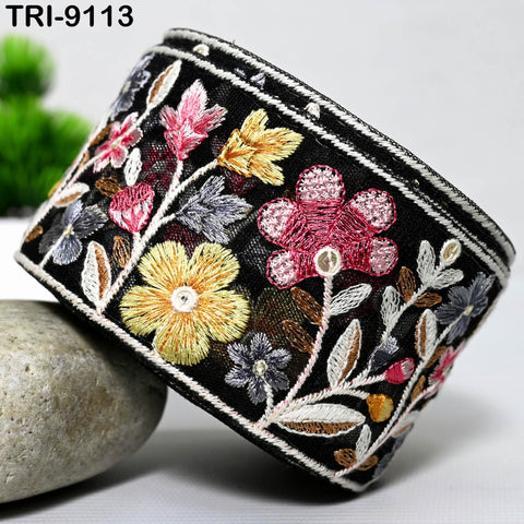 a decorative black and white fabric with a floral pattern, including yellow, pink, and white flowers, as well as leaves and stems. The fabric appears to be a piece of clothing or accessory with a detailed embroidered design.