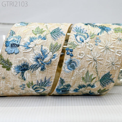a close-up view of a decorative fabric with a floral pattern, featuring blue and white flowers and leaves. The fabric appears to be a beige color, and the intricate embroidery details are visible, creating a visually appealing and textured design.