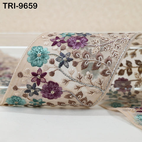 a decorative fabric with a floral pattern, including purple and blue flowers, as well as leaves and stems. The fabric appears to be a lace or sheer material, with a beige or off-white background.
