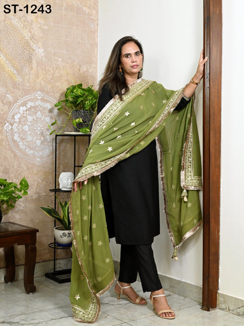 A woman wearing a green and gold patterned shawl stands in a room with a wooden door and a plant on a shelf.