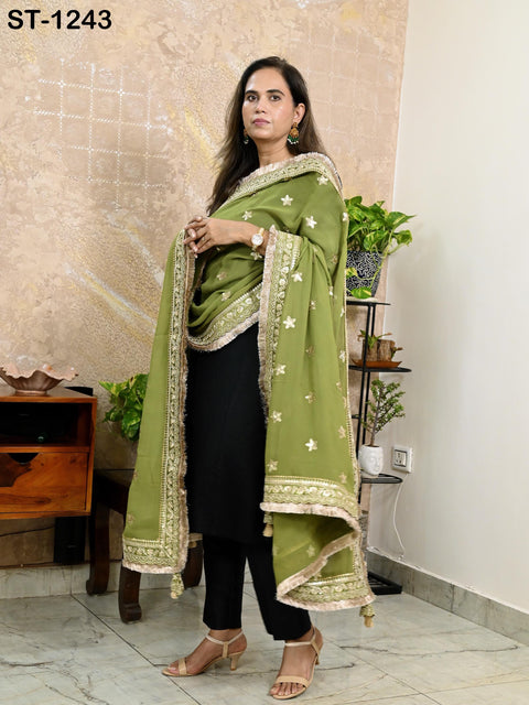 A woman wearing a green and gold shawl and black pants stands in a room with a wooden dresser and potted plants.
