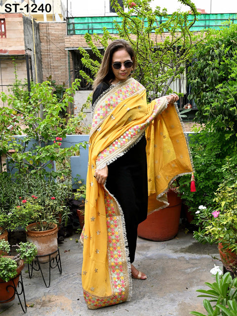 A woman wearing a yellow and orange patterned shawl stands in a garden surrounded by potted plants.