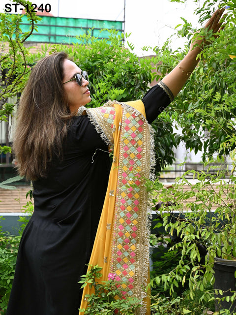 A woman in a black dress reaches up to touch leaves on a tree, wearing sunglasses and a colorful shawl.