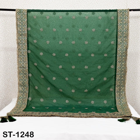 a green and gold embroidered fabric with a repeating pattern of small white flowers or dots. The fabric is displayed on a white surface, possibly a table or a stand, with a black frame around it. The image does not contain any text or additional objects.