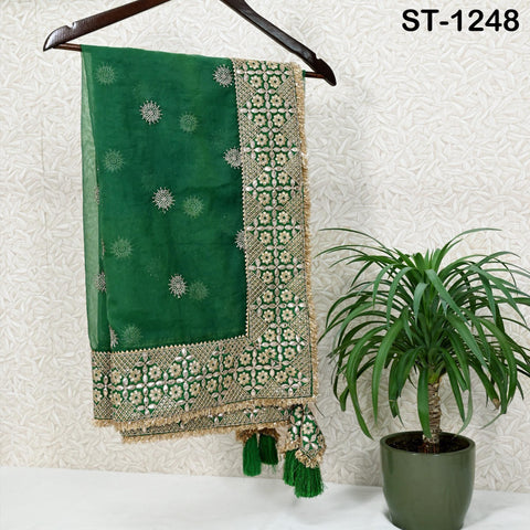 a green and gold embroidered fabric hanging on a wooden hanger, placed next to a potted plant.