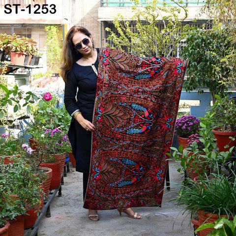 A woman in a black dress stands in a garden surrounded by potted plants, holding up a colorful, patterned fabric.