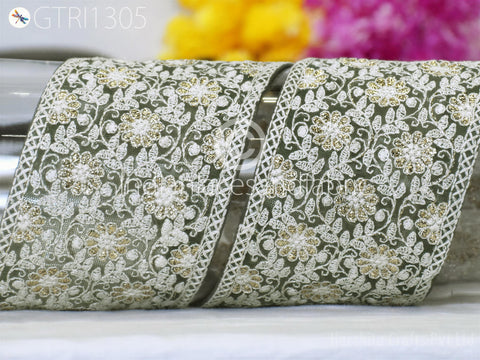 a close-up view of a decorative fabric with a floral pattern, including white and gold elements. The fabric appears to be a lace or embroidered material, with a repeating floral design. The background is blurred, but it seems to be a colorful floral arrangement.