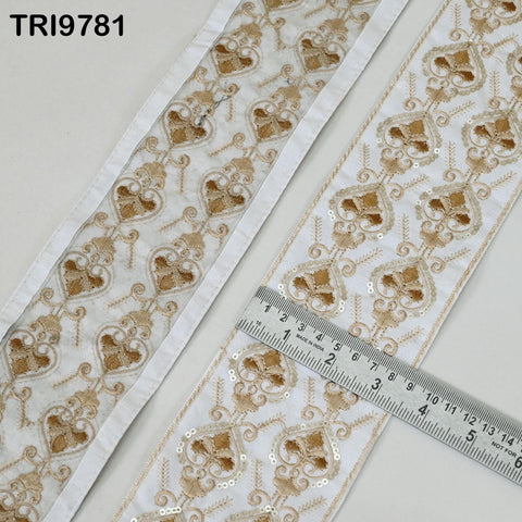 a close-up view of a decorative fabric with a repeating pattern of gold and white floral designs. The fabric appears to be made of a material with sequins, and a ruler is placed on top of it to measure the length.