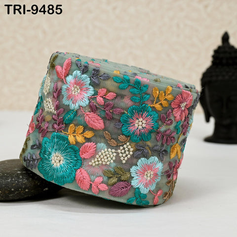 a decorative fabric with a floral pattern, including various shades of pink, blue, green, and yellow flowers, as well as leaves and beads. The fabric appears to be a piece of clothing or accessory with intricate embroidery.