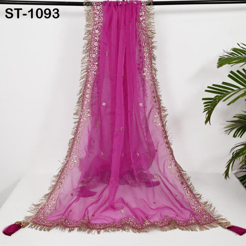 a long, pink, sheer fabric with a gold and pink fringe trim. The fabric appears to be a sheer chiffon or sheer curtain, with a decorative border. The background is a plain white wall, and there is a plant visible on the right side of the image.