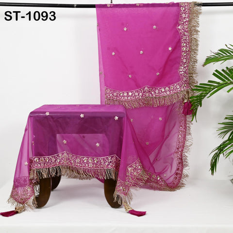 a pink and gold decorative fabric with a floral pattern, hanging on a black rod. The fabric appears to be a tablecloth or a decorative piece, possibly for a table or a table runner. The background is a plain white wall, and there is a potted plant visible on the right side of the image.