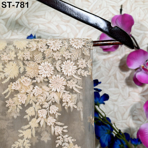 a close-up view of a delicate, lace-like fabric with intricate floral embroidery. The fabric is hanging on a wooden hanger, and there are purple flowers visible in the background.