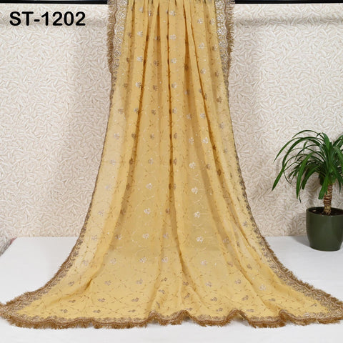 a long, yellow, embroidered shawl with a gold trim, hanging on a wall. Next to the shawl, there is a potted plant, creating a decorative and cozy atmosphere.