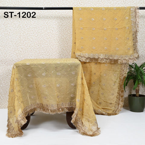 a pair of yellow, intricately embroidered fabrics hanging on a black rod. The fabrics are adorned with gold and white embroidery, adding a touch of elegance to the scene. The background includes a white wall with a floral pattern, and a potted plant is visible on the right side of the image.