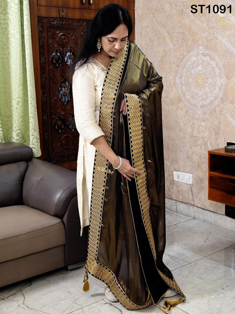 A woman in a white and gold dress stands in a room, holding a black and gold shawl.