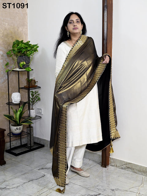 A woman wearing a white dress with a gold and black patterned shawl stands in a room with a potted plant and a shelf with decorative items.