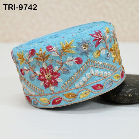 A close-up image of a decorative blue fabric with intricate floral embroidery, including red, yellow, and white flowers, along with gold and blue accents.