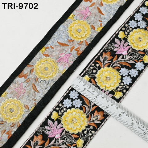 two pieces of fabric with intricate floral embroidery. One piece is black and the other is gray, both featuring a variety of yellow and pink flowers. A ruler is placed on the gray fabric, likely for measuring the length or width of the fabric.